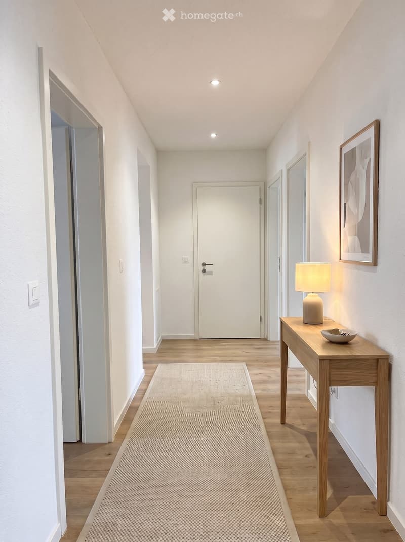 Modern hallway with wooden flooring, white walls, a small wooden table with a lamp and bowl, and a framed picture on the wall.