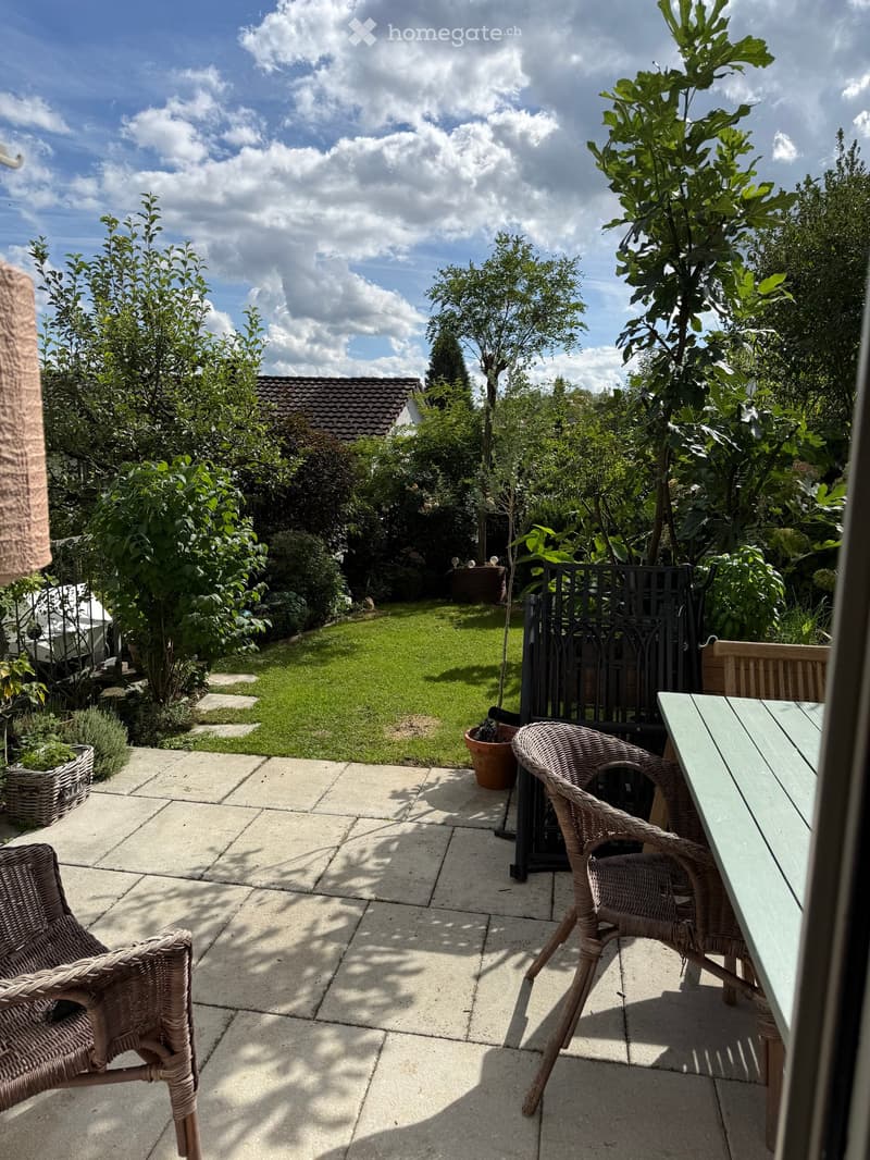 A well-maintained garden with a paved patio area featuring wicker chairs and a table, surrounded by lush greenery and trees.