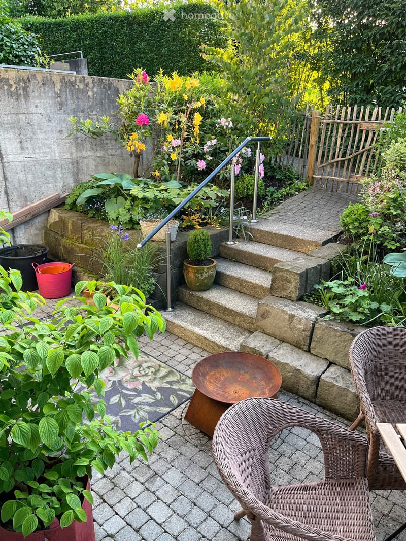 A well-maintained garden area with a stone-paved patio, featuring wicker chairs, a small table, potted plants, and colorful flowers. A handrail leads up a set of stone steps.
