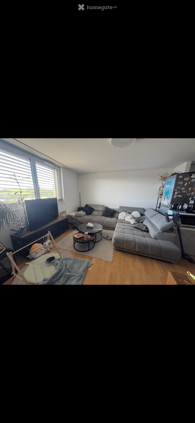 Modern living room with grey sectional sofa, coffee table, TV stand, and a small table with chairs. The room features wooden flooring, large windows with blinds, and a cozy rug.
