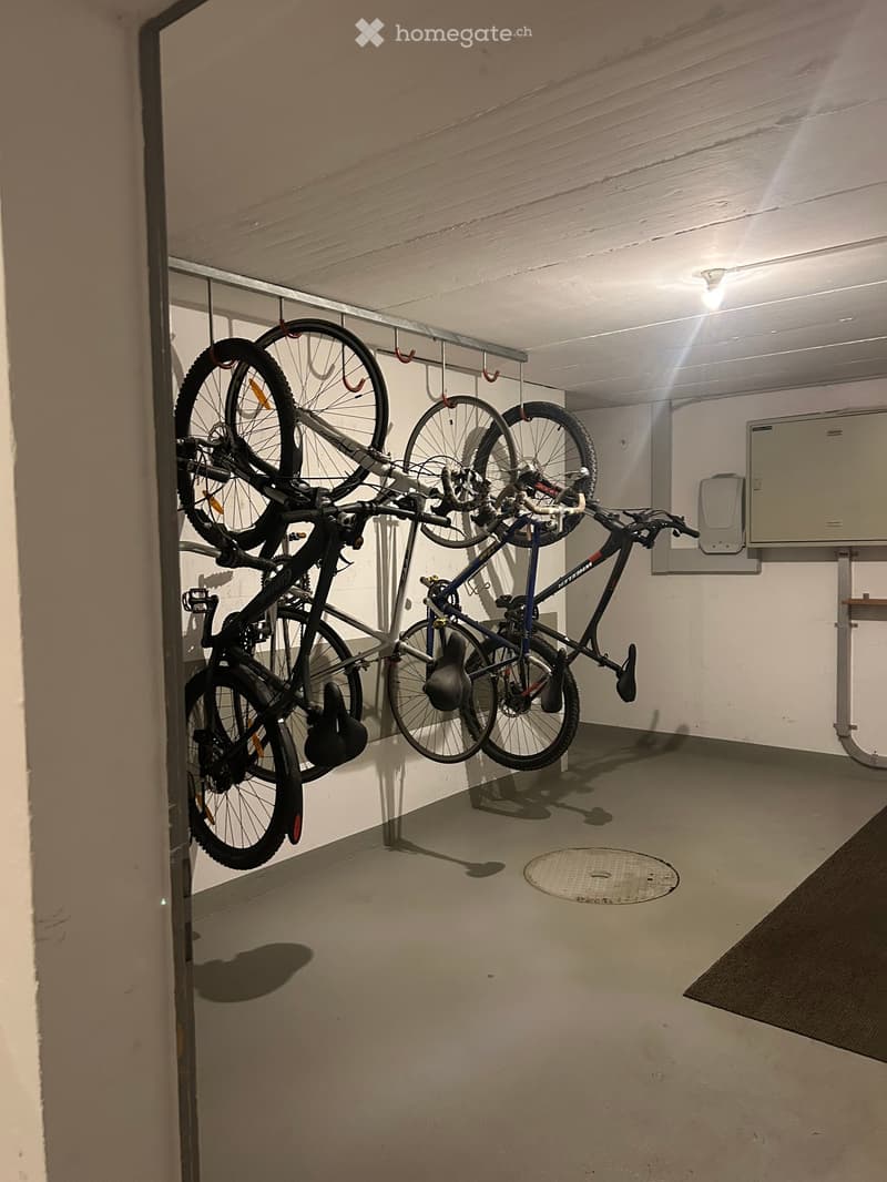 A small, well-lit storage area with several bicycles hanging from a rack, helmets placed on the floor, and a clean, concrete floor.