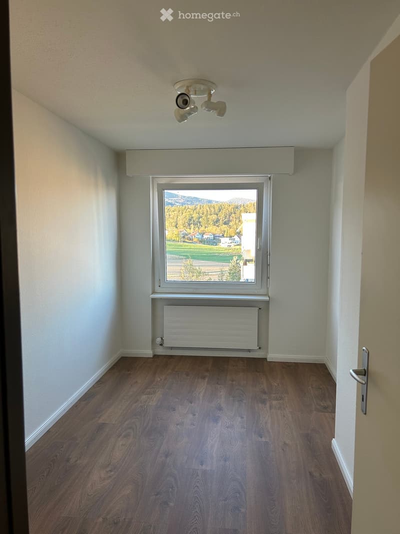 Empty room with wooden flooring, white walls, a large window with a view of trees and a radiator below it.