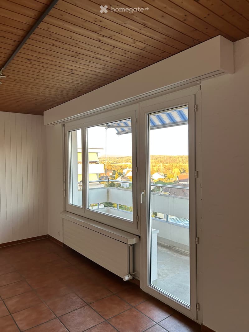 Empty room with wooden ceiling, tiled floor, radiator, and large window opening to a balcony with a view of the landscape.