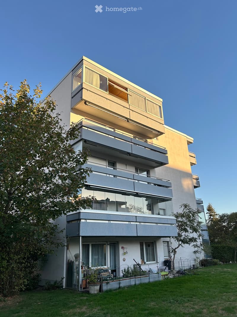 Modern multi-story apartment building with balconies, large windows, and a landscaped garden area in front.