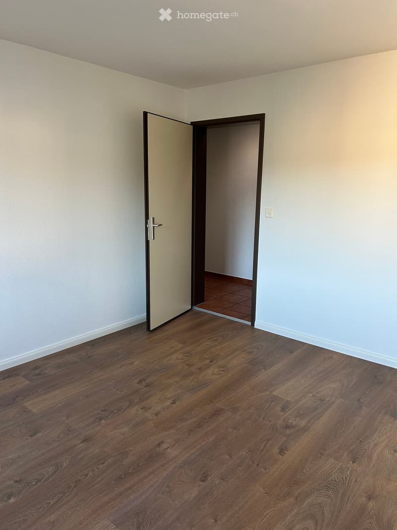 Empty room with wooden flooring and white walls, featuring an open door leading to another room with tiled flooring.