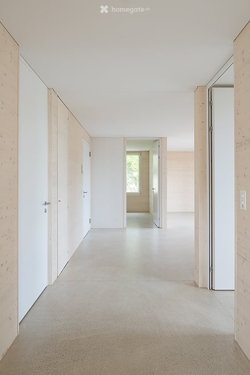 Empty hallway with white walls, light wooden paneling, and a smooth concrete floor. The hallway leads to an open doorway revealing a glimpse of another room with a window and light-colored walls.