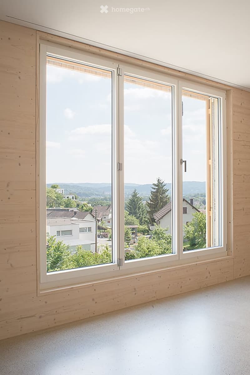 Empty room with wooden walls and large windows offering a scenic view of the landscape and neighboring houses.