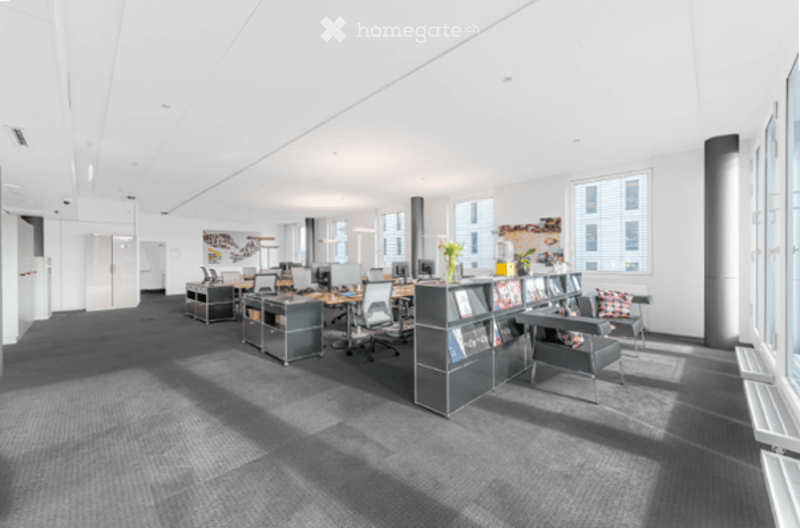 Modern office space with desks, chairs, shelves, and a small seating area with a chair and a flower vase.