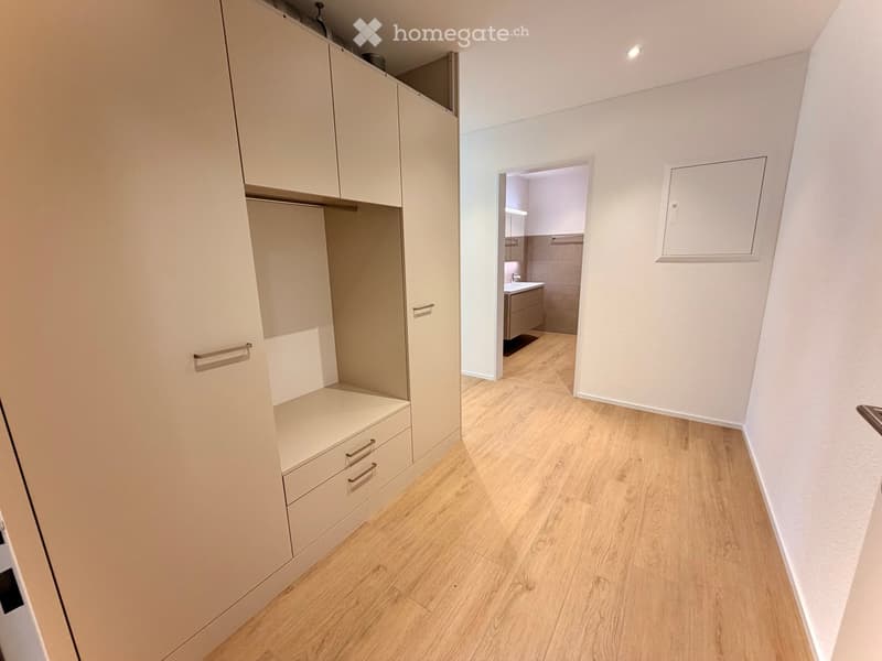A clean, modern hallway with light wooden flooring and white walls. A built-in wardrobe with drawers and shelves is on the left, and an open doorway leads to a small bathroom with a sink and tiled walls.