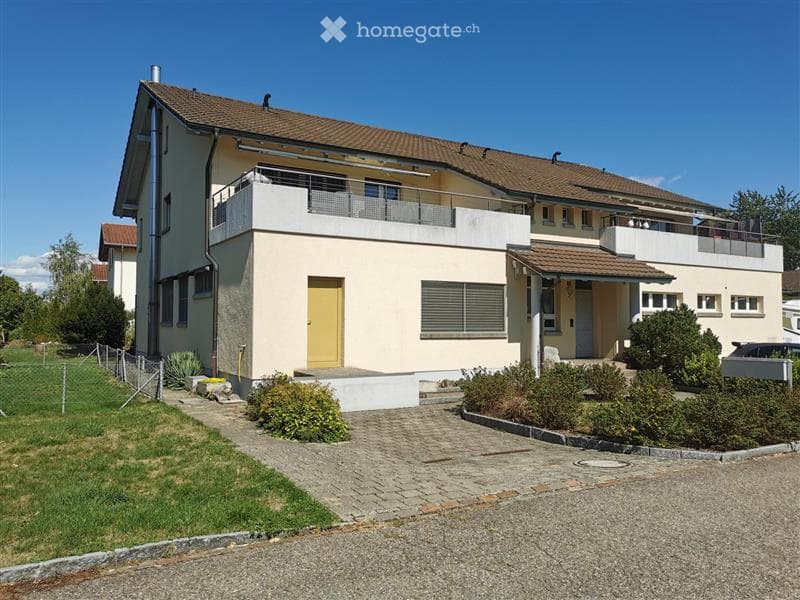 Two-story house with a beige facade, brown tiled roof, and multiple balconies. The house features a yellow front door, multiple windows, and a well-maintained garden with shrubs and a paved driveway.