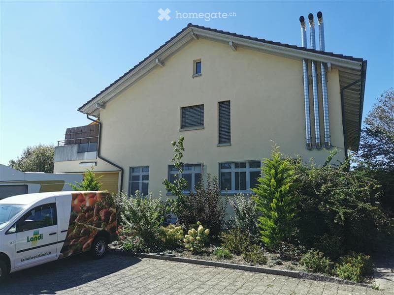 Single-family house with a light-colored facade, multiple windows, and a small garden in front.