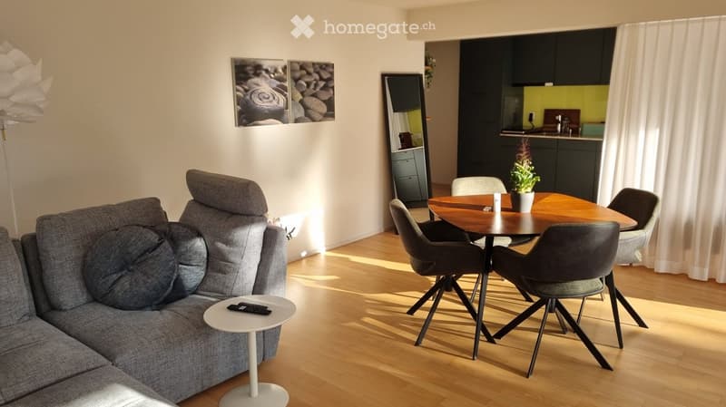 Modern living and dining area with a grey sofa, coffee table, dining table with chairs, and a potted plant. The space features wooden flooring and large windows with white curtains.