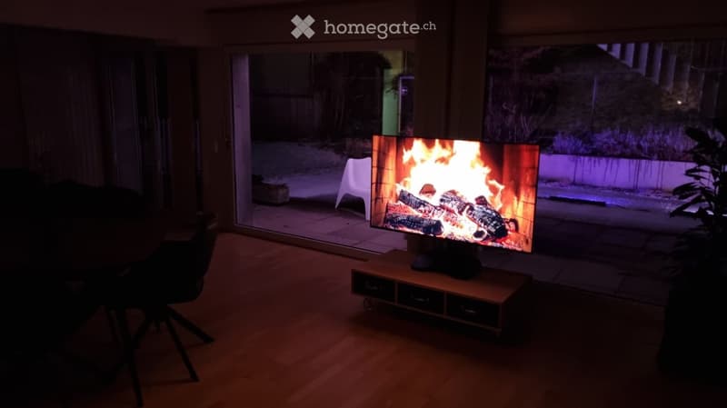 Living room with a large television displaying a fireplace scene, TV stand, and a white chair near the sliding glass door leading to an outdoor area.