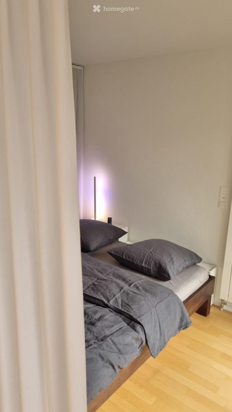 A small, neatly arranged bedroom with a single bed, dark grey bedding, two pillows, a wooden nightstand, and a tall, slim lamp.