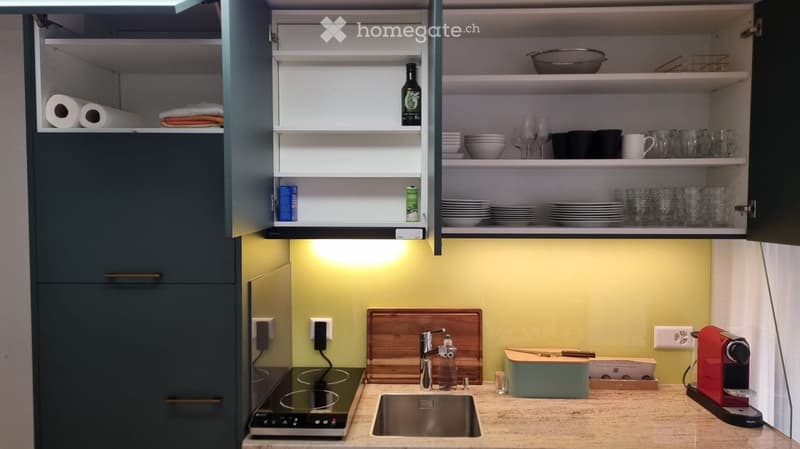 Modern kitchen with dark grey cabinets, a light yellow accent wall, and a beige countertop. Features include a sink with a modern faucet, a coffee maker, a wooden cutting board, and various kitchen items on the shelves and countertop.