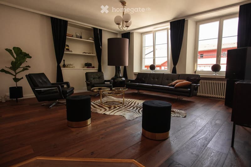 Modern living room with wooden flooring, large windows, a black leather sofa, two black leather chairs, two round gold tables, two black stools, a large floor lamp, a plant, and a shelf with books and decorative items.
