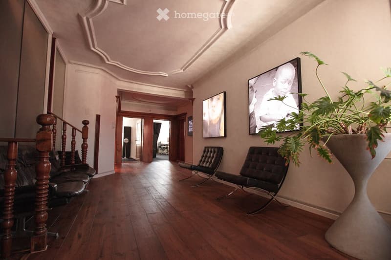 Elegant living room with wooden flooring, a staircase with wooden balusters, two black leather chairs, a large plant in a white pot, and two picture frames on the wall.