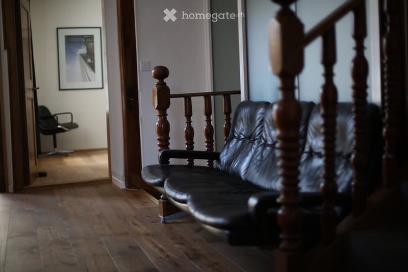 A furnished room with a black leather sofa and a black office chair, wooden flooring, and a staircase with wooden railings.