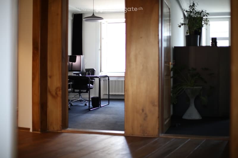 Office space with a desk, chair, computer, and potted plant, viewed through an open wooden door.