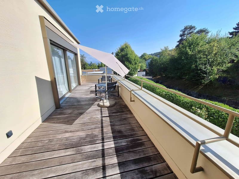 Wooden balcony with a table, chairs, and an umbrella, overlooking a green, tree-lined area.