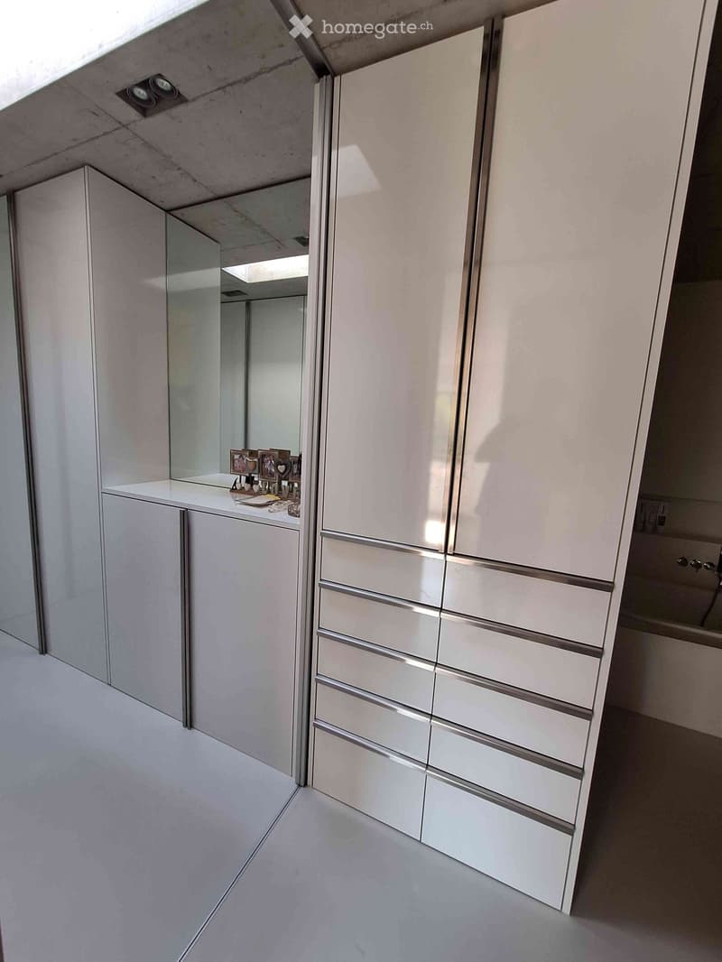 Modern, minimalist walk-in closet with white cabinets, a large mirror, and a small table holding framed photos and decorative items.