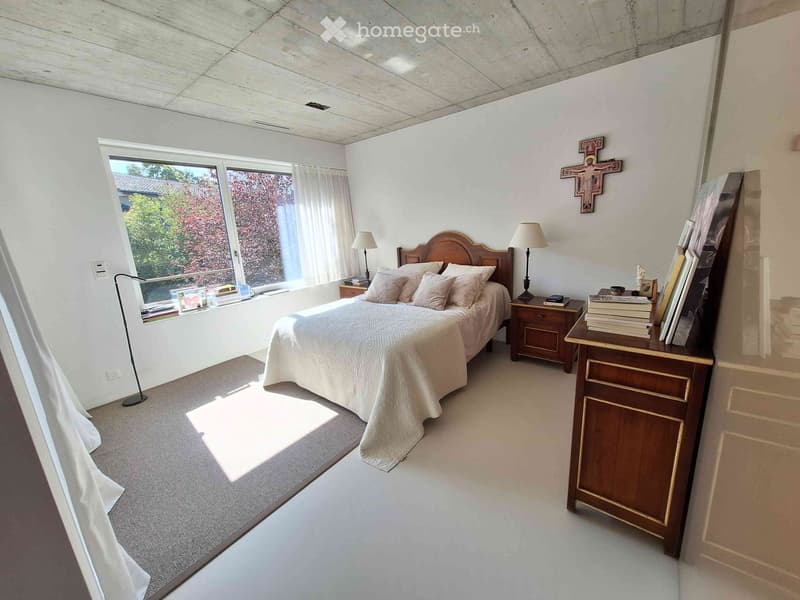 Bright bedroom with a large window, a wooden bed with a beige comforter, nightstands with lamps, a wooden dresser with books, and a cross on the wall.
