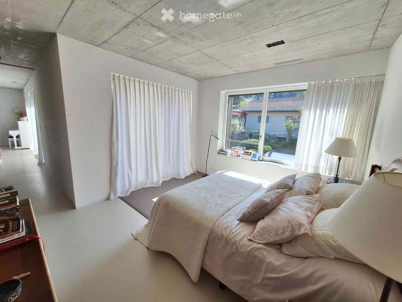 Modern bedroom with a large bed, nightstand, lamp, and large window with curtains. A small table and chair are visible in the background.