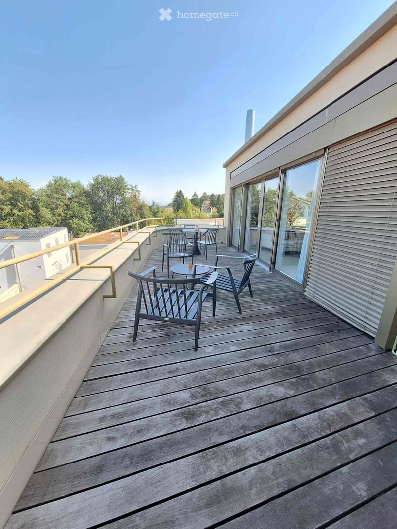 Wooden deck with modern chairs and tables, overlooking a green area with trees.