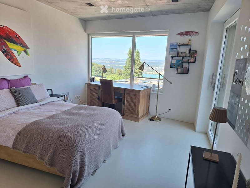 Bright bedroom with a large window offering a scenic view, featuring a bed with pillows and a blanket, a wooden desk with a chair, and various lamps and decor.
