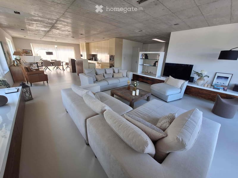 Modern living room with white walls, large windows, and a concrete ceiling. Features include a large sectional sofa, coffee table, armchairs, dining area with a table and chairs, and a built-in entertainment unit with a TV and shelves.