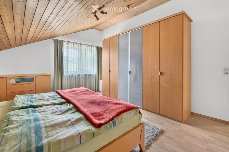 Cozy bedroom with wooden bed, plaid blanket, large wardrobe, dresser, and window with curtains.
