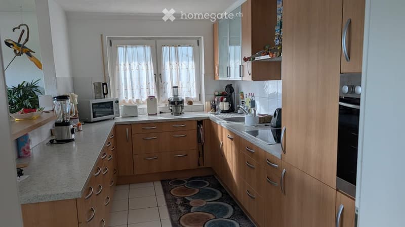 A modern kitchen with wooden cabinets, granite countertops, and various appliances including a microwave, blender, and coffee maker. The space is well-lit with natural light from a window and features a tiled floor and a decorative rug.