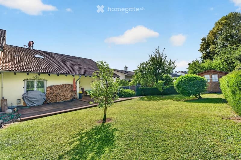 Well-maintained garden with a wooden deck, covered grill, stack of firewood, small table and chairs, and a small shed.