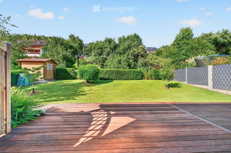 A well-maintained garden with a wooden deck, green lawn, trimmed hedges, and a small wooden shed. The area is surrounded by trees and a modern fence.