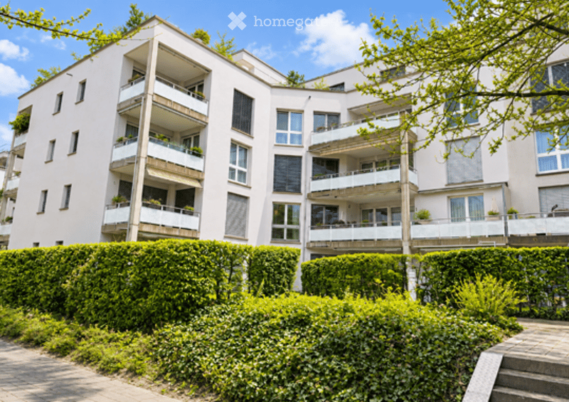 Modern multi-story apartment building with white facade, multiple balconies, and greenery in front.