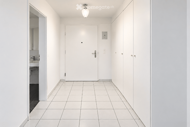 A clean, white-tiled hallway with a single closed door, a light fixture, and a small adjacent bathroom visible on the left side.