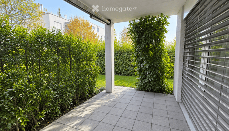 A modern outdoor patio with tiled flooring, surrounded by lush green shrubs and trees, with a covered walkway leading to a building.