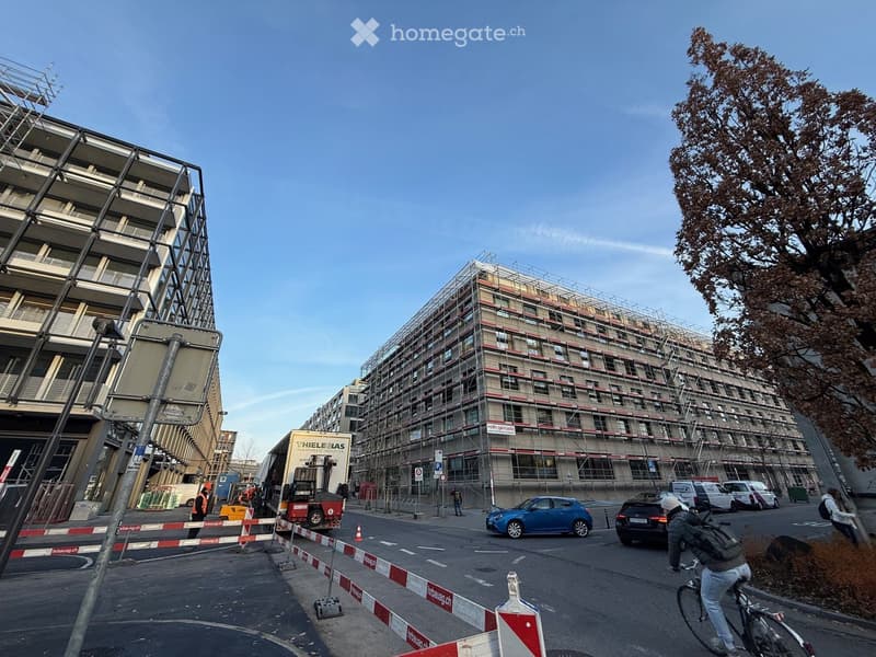 A street with a blue car and a cyclist, construction trucks and scaffolding on a large building, another building in the background, and people in vests and hats standing by a barrier