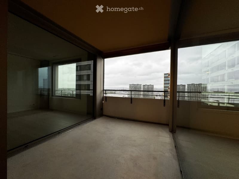 Empty balcony with large windows offering a cityscape view, carpeted floor, and metal railing.