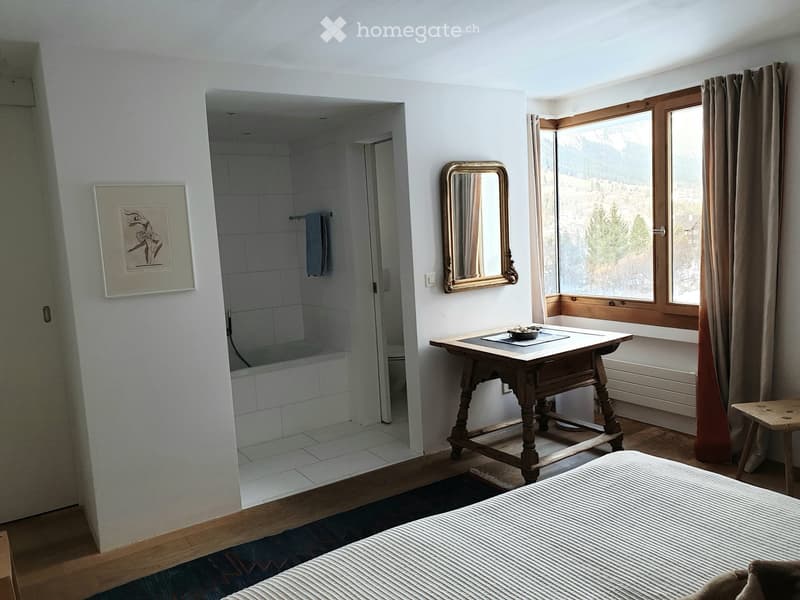 Cozy bedroom with a wooden bed, small wooden table, mirror, and an adjacent bathroom with a bathtub.