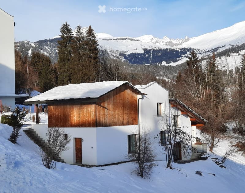 Modern white house with wooden accents, snow-covered roof, and steps leading to the entrance, surrounded by snow-covered trees and mountains.