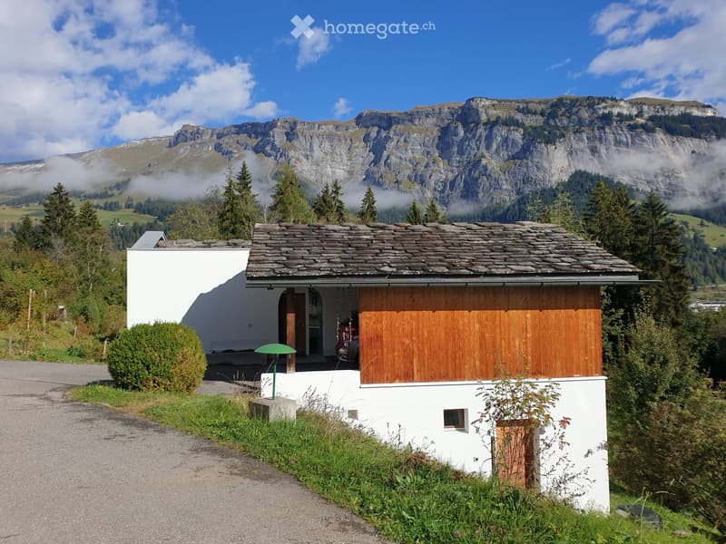 A small, modern white house with a stone roof, surrounded by lush greenery and mountains in the background.
