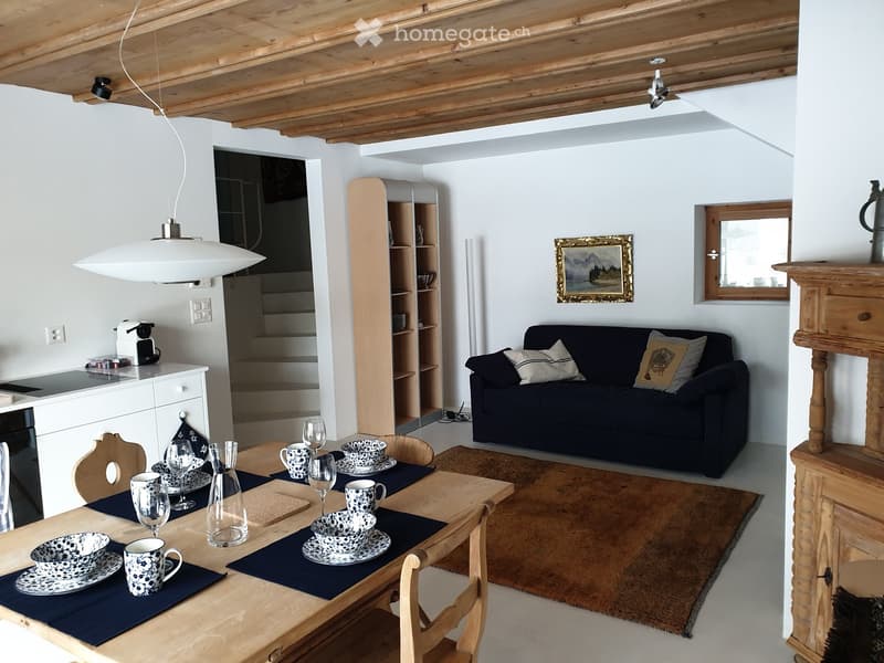 Open-plan living and dining area with exposed wooden beams, a dark blue sofa, a wooden dining table set with cups and glasses, and a pendant light hanging from the ceiling.