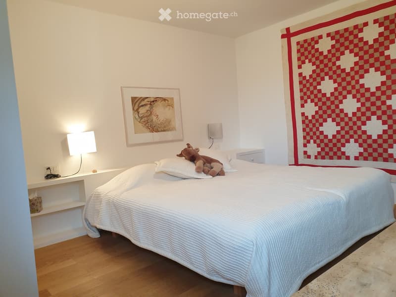 Modern bedroom with a large bed, white bedding, a nightstand with a lamp, a framed picture on the wall, and a decorative quilt on the bed.