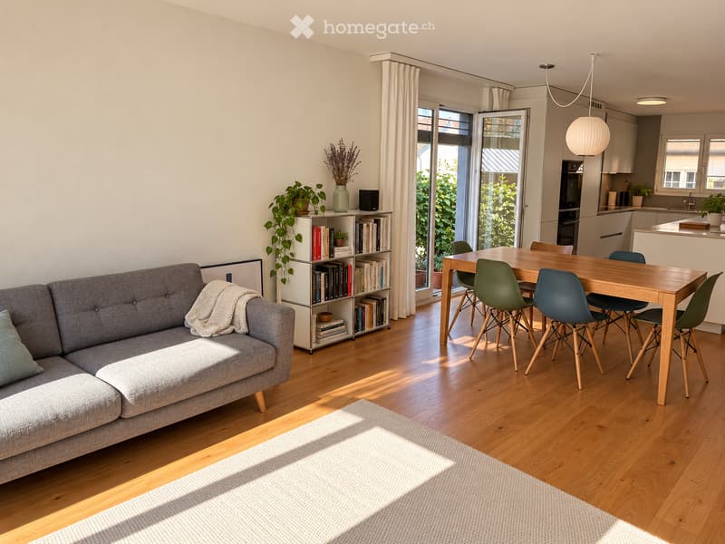Bright living room with grey sofa, wooden dining table, chairs, bookshelf, and plants. Natural light from large windows and sliding doors.