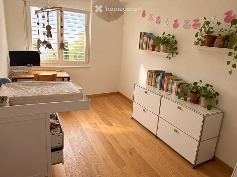 A well-lit children's room with a lofted bed, desk, shelves filled with books and plants, and a hanging toy mobile above the bed.