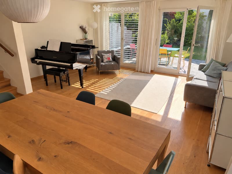 Bright living room with wooden flooring, large windows, and sliding glass doors leading to a patio. Features a grey sofa, wooden dining table with chairs, a grand piano, and a floor lamp.