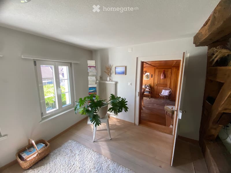 Bright hallway with wooden flooring, white walls, a large window, a small table with a potted plant, a radiator, a picture frame, a wicker basket, and an open doorway leading to another room with wooden furniture.