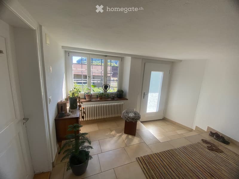 Hallway with tiled floor, white walls, large window with potted plants on sill, small table with items, radiator below window, door leading outside, small nest decoration, striped rug, and shoes near entrance.