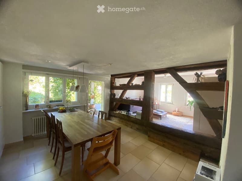 Bright dining area with a wooden table and chairs, shelves with decorative items, and large windows letting in natural light.